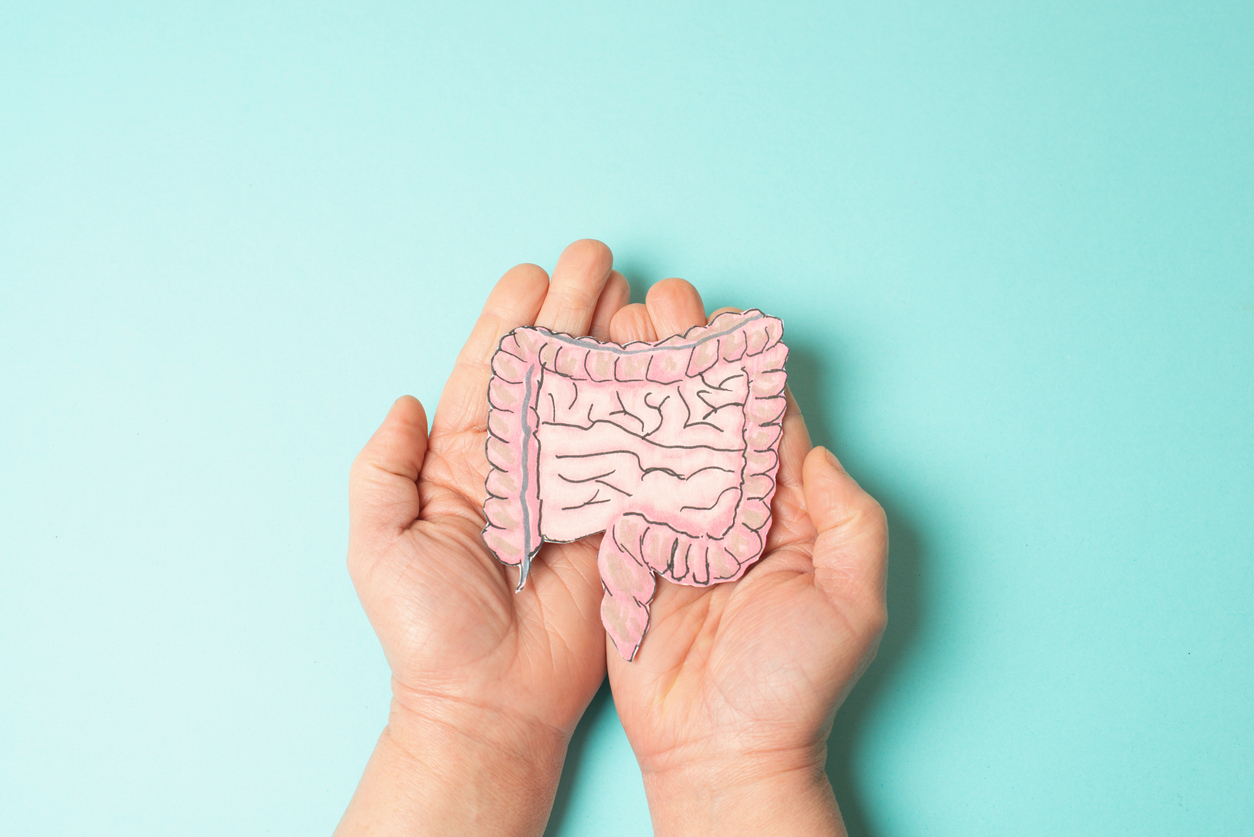Hands gently holding a paper cutout illustration of the human large intestine and rectum on a teal background, representing bowel health care.